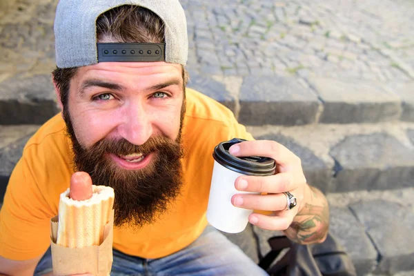 Conceito de cultura alimentar urbana. Hipster comer cachorro-quente enquanto se sentar em escadas ao ar livre. Hipster desfrutar de cachorro quente e beber xícara de papel. Homem barbudo desfrutar de comida de rua fundo urbano. Faça uma pausa para comer um lanche — Fotografia de Stock