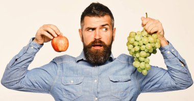 Sakallı adam bir demet yeşil üzüm ve elma tutuyor.