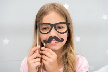 Kız, yatak odasında fotoğraf kabini aksesuarları gözlükler ve bıyıkla poz veriyor. Parti gözlüklü kız çocuğu. Çocuk erkekliği taklit eder ya da babasına benzemek ister. Bıyıklı ve gözlüklü çocuk.