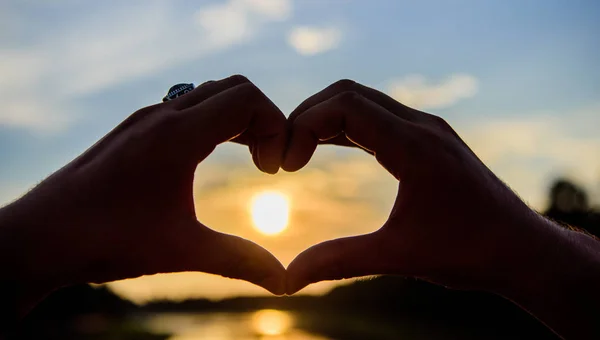 Mãos masculinas em forma de coração gesto símbolo de amor e romance. gesto do coração em frente ao pôr do sol acima da superfície da água do rio, desfocado. Os melhores lugares para encontros românticos. Pôr do sol atmosfera romântica — Fotografia de Stock