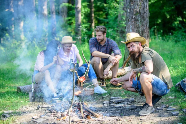 Hipster asando salchicha mientras sus amigos hablan compartiendo impresión y viendo fotos en cámara. Turistas tomando un aperitivo con comida asada sobre fuego. Amigos grupo turístico relajarse cerca de la hoguera — Foto de Stock