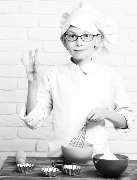 Jeune garçon petit chef cuisinier mignon en uniforme blanc et chapeau sur le visage souriant avec des lunettes debout près de la table avec rouleau à pâtisserie et bols colorés et cuisine sur fond de mur de briques — Photo