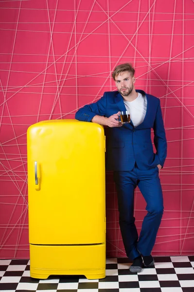 Man formal elegant suit lean on retro vintage yellow refrigerator. Bachelor elegant guy drink tea or coffee near fridge. Bright fridge household appliances interior object. Morning coffee concept