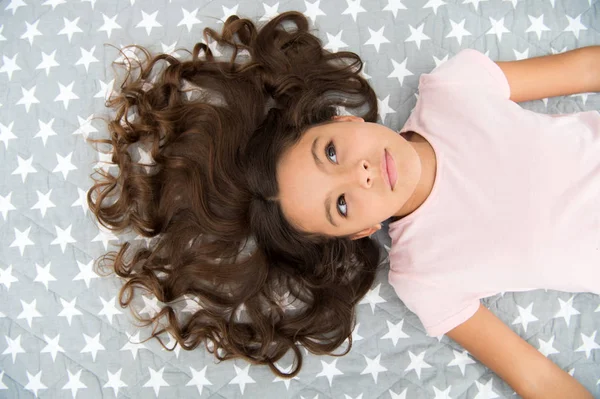 長い巻き毛の女の子の子供はベッドの上のビューに横たわっていた。子供の完璧な巻き毛はかわいいように見えます。コンディショナーマスク有機オイルは、髪の光沢と健康を維持します。驚くべきカールのヒント。巻きつけるのは自然だけど — ストック写真