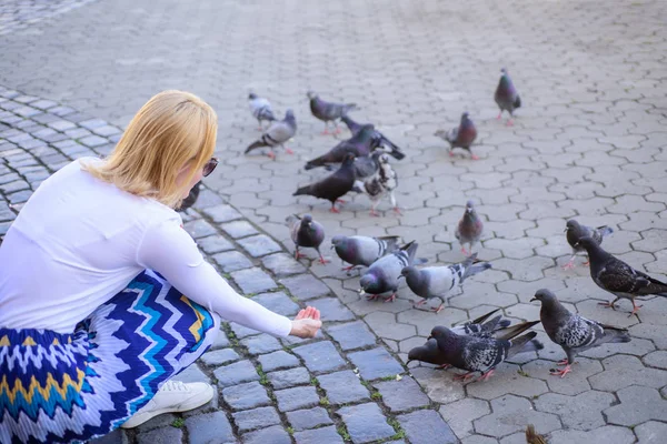 Touristinnen oder Bürger werfen Krümel für Tauben. Mädchen blonde Frau entspannen Stadtplatz und Tauben füttern. Mädchen füttert Taubenvögel. Tauben warten auf dem Stadtplatz auf Leckereien. Großzügigkeit teilen — Stockfoto