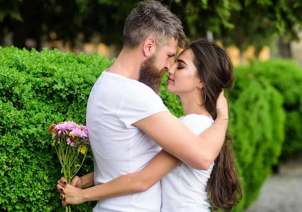 Le gusta su hermoso cabello. Chica sostiene flores mientras el hombre acaricia su pelo largo. Hombre barbudo hipster abraza suavemente novia disfrutando de su pelo largo suave. Pareja en amor abrazos al aire libre parque fondo — Foto de Stock