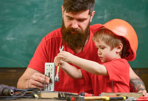 Rapaz, criança ocupada com capacete protector faz à mão, reparar, faz artesanato com o pai. Mens conceito de trabalho. Pai com barba ensinando o filhinho a usar ferramentas na sala de aula, quadro no fundo — Fotografia de Stock