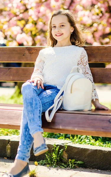 Menina no rosto sorridente senta-se no banco, árvore sakura no fundo, desfocado. Menina relaxante durante o passeio no parque perto de flor de cereja. Relaxe o conceito. Criança bonito com saco ou mochila desfrutar de dia de primavera ensolarado — Fotografia de Stock