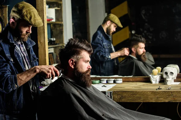 Barber with hair clipper works on hairstyle for man with beard, barbershop background. Barber styling hair of bearded client with comb and clipper. Hipster client getting haircut. Haircut concept