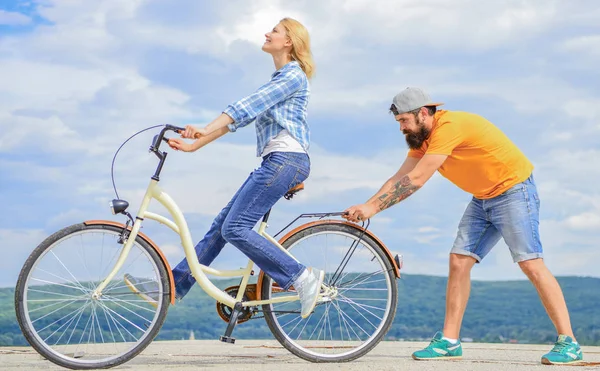 How to learn to ride bike as an adult. Girl cycling while boyfriend support her. Teach adult to ride bike. Man helps keep balance and ride bike. Find balance. Woman rides bicycle sky background
