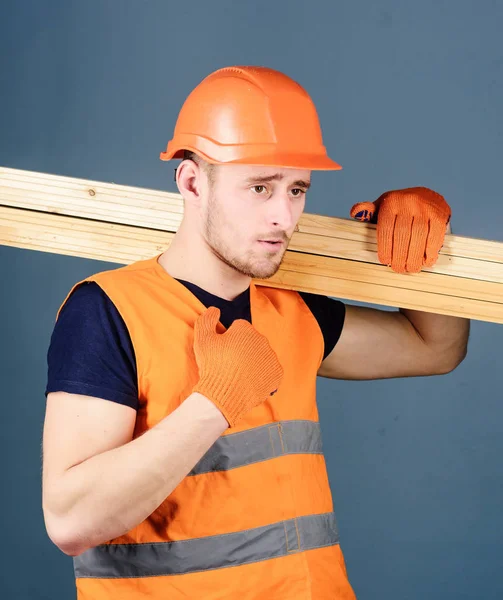 Carpenter, woodworker, strong builder on serious face carries wooden beam on shoulder. Wooden materials concept. Man in helmet, hard hat and protective gloves holds wooden beam, grey background.