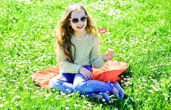 草地、緑の背景に芝生の上に女の子が座っています。陽気な顔の女の子は、アウトドア レジャーをお過ごしください。ファッショニスタのコンセプトです。子供サングラス段ボール草原でのフォト セッションの唇を笑顔でポーズ. — ストック写真
