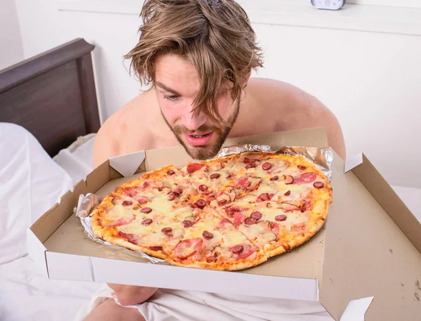 Man likes pizza for breakfast. Who cares about diet. Man bearded handsome guy eating cheesy food for breakfast in bed. Guy holds pizza box sit bed in bedroom or hotel room. Food delivery service.