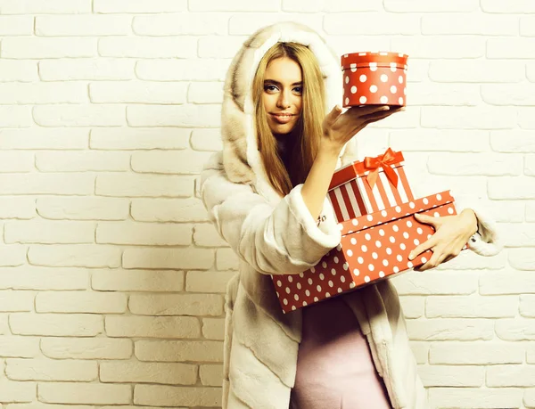 Jeune femme sexy jolie à la mode ou fille avec de longs beaux cheveux blonds en manteau de fourrure blanche avec capuche et maquillage de mode tenant de grands cadeaux rouges ou cadeaux sur fond de studio de mur de brique — Photo