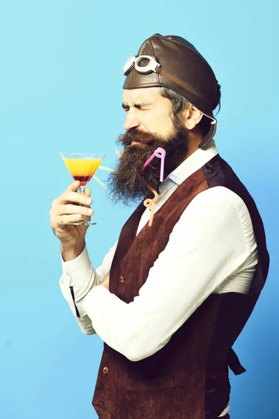 handsome bearded aviator man with long beard and mustache on displeased face holding glass of alcoholic beverage in vintage suede leather waistcoat with hat and glasses on red studio background.
