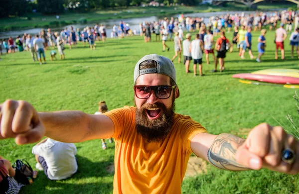 Cheerful fan at summer fest. Man bearded hipster in front of crowd people raise fists green riverside background. Hipster in cap happy celebrate event picnic fest or festival. Urban event celebration