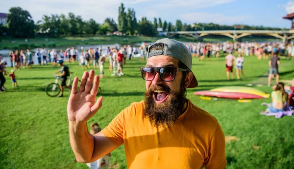 Happy to meet you. Urban event celebration. Hipster in cap happy to meet friend at event picnic fest or festival. Man bearded hipster in front of crowd people waving hand green riverside background