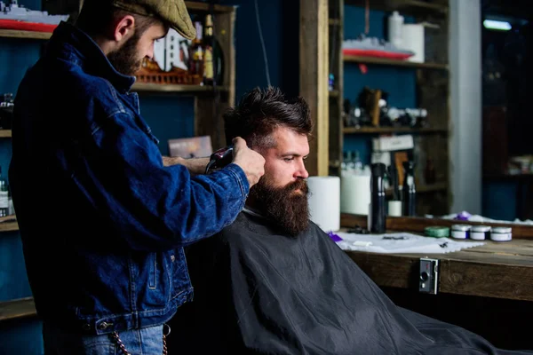 Um cliente Hipster a cortar o cabelo. Barbeiro com cortador de cabelo funciona no penteado para barbearia barba cara fundo. Conceito de estilo de vida Hipster. Barbeiro com clipper aparar o cabelo no templo do cliente — Fotografia de Stock