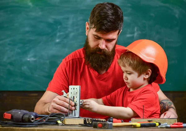 Father with beard and little son in classroom teaching to use tools, chalkboard on background. Boy, child in protective helmet makes by hand, repairing, does crafts with dad. Mens work concept