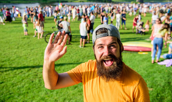 Человек бородатый хипстер перед толпой людей зеленый фон поля. Хипстер в кепке посещает пикник или фестиваль. Празднование городского события. Человек машет рукой солнечный день на открытом воздухе
.