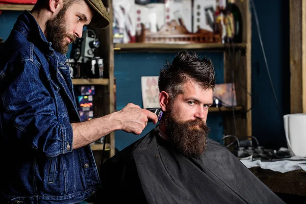 Concetto di stile di vita hipster. Il barbiere con tagliacapelli lavora sull'acconciatura per lo sfondo del barbiere. Barbiere con taglierina che taglia capelli su tempio di cliente. cliente hipster ottenere taglio di capelli — Foto Stock