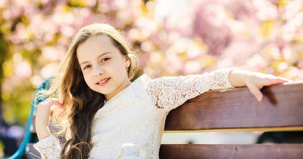 Conceito de beleza natural. Menina no rosto sorridente senta-se no banco, árvore sakura no fundo, desfocado. Criança bonito com cabelo longo e bonito desfrutar de dia de primavera ensolarado. Menina relaxante no parque perto de flor de cereja — Fotografia de Stock