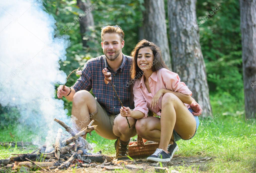 Couple in love enjoy camping forest roasting sausage at bonfire. Camping and picnic. Traditional roasted food as attribute of picnic. Couple roasting sausages on sticks nature background