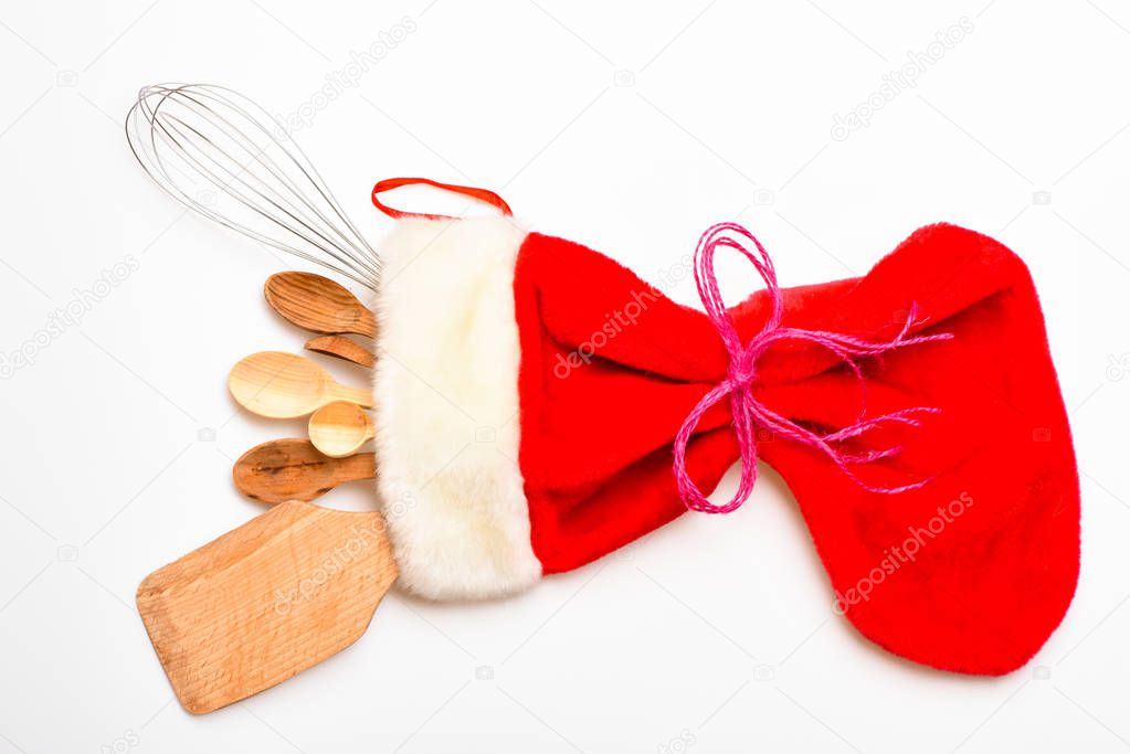 Christmas gift concept. Kitchen accessories or kit of kitchenware packed in big red sock, white background. Kitchenware as wooden spoons and whisk lay in Christmas sock. Kitchen accessories or tools