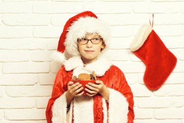 Dekoratif Noel veya Noel çorap veya çikolata kalça kurabiye kase beyaz tuğla duvar arka planda tutan önyükleme ile kırmızı kazak ve yeni yıl şapkalı gözlüklü Genç Sevimli Noel Baba Çocuk