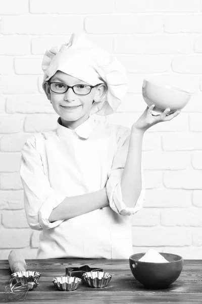 흰색 유니폼 소년 작은 귀여운 쿡 요리사와 웃는 얼굴이 케이크와 벽돌 벽 바탕에 청록색 지주 그릇에 대 한 롤링 핀 금형 테이블 근처에 서 안경에 모자 — 스톡 사진