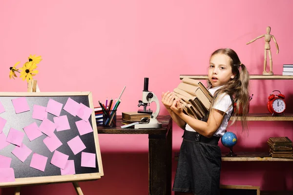 Dívka nese hromadu knih na stůl s mikroskopem — Stock fotografie