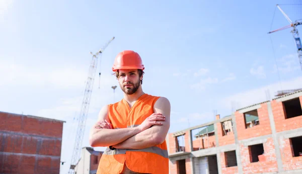 Concepto de responsabilidades del contratista. Un tipo con casco protector parado frente a un edificio hecho de ladrillos rojos. Proceso de construcción de control. Hombre en chaleco naranja y obras de casco en el sitio de construcción — Foto de Stock