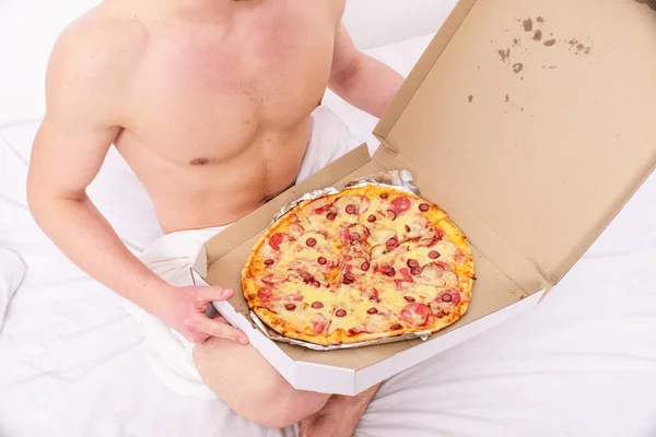 Courier masculin sexy torse musculaire tient pizza. Courrier sexy offre une satisfaction gastronomique à votre lit. Guy nu couvert boîte à pizza assis lit chambre à coucher vous offrent de le rejoindre. Satisfaction gastronomique — Photo