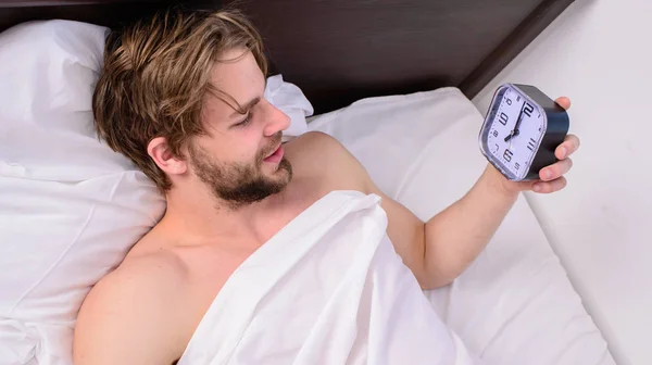Stick sleep schedule same bedtime and wake up time. Sleep regime habits concept. Man sleepy drowsy unshaven bearded face covered with blanket having rest. Man unshaven lay awake bed hold alarm clock — Stock Photo, Image