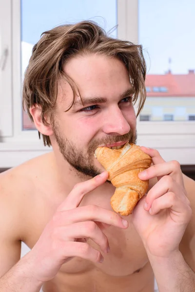 Goditi ogni boccone. L'uomo mangia croissant gli piacciono i prodotti da forno. Il tipo morde il dolce dolce. Un bel tipo barbuto che fa colazione a letto. Guy tiene croissant camera da letto camera d'albergo finestra sfondo — Foto Stock