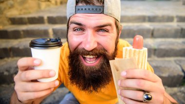 Hipster sosisli sandviç içki kağıt bardak tadını çıkarın. Sakallı adam hızlı öğle yemeği merdiven arka plan tadını çıkarın. Fast food yemek yemek için. Sokak gıda diyet için önemli bir katkı yapmak. Hipster sosisli sandviç yemek