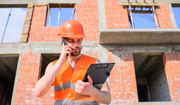 Guy protective helmet stand in front of building made out of red bricks. Builder vest and helmet works at construction site. Contractor control according to plan. Engineer phone call control process