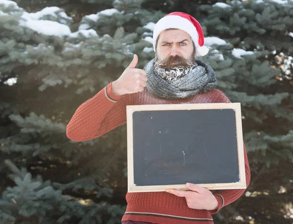 Frons man in santa claus hoed, hipster met baard en snor bedekt met witte vorst, met blanco zwart bord of krijtbord geeft duimen omhoog gebaar op winterdag op natuurlijke achtergrond, kopieer ruimte. — Stockfoto