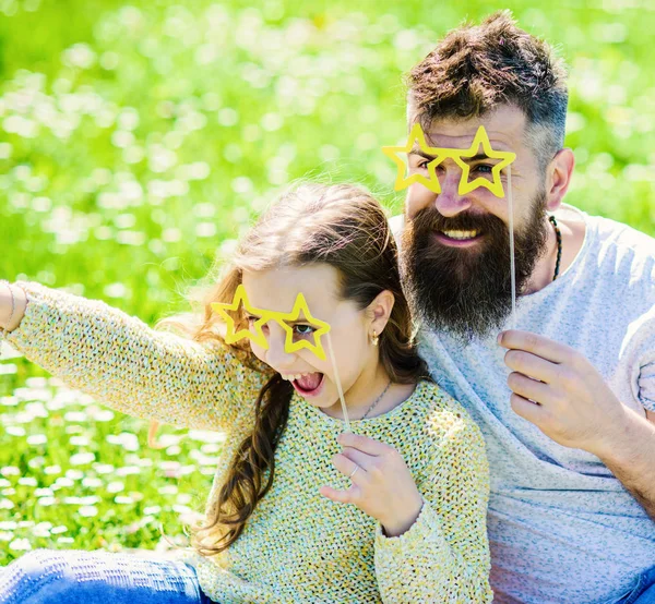 Dad and daughter sits on grass at grassplot, green background. Child and father posing with star shaped eyeglases photo booth attribute at meadow. Star and fame concept. Family spend leisure outdoors — Stock Photo, Image