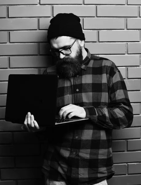 Homem barbudo, barba comprida. Brutal caucasiano grave sem barba hipster segurando laptop em vermelho camisa xadrez preto com chapéu e óculos no fundo do estúdio de parede de tijolo marrom. — Fotografia de Stock