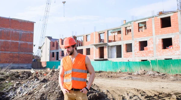 Homme en gilet orange et casque travaille sur le chantier de construction. Concept des responsabilités de l'entrepreneur. Le type au casque de protection se tient devant le bâtiment en briques rouges. Processus de construction de contrôle — Photo