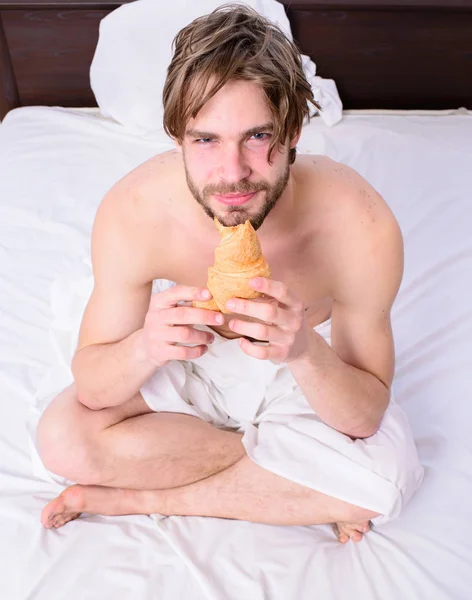 Man eats croissant he likes bakery products. Fresh bakery product. Man bearded handsome guy eating breakfast in bed. French breakfast stereotype. Guy holds croissant sit bed in bedroom or hotel room