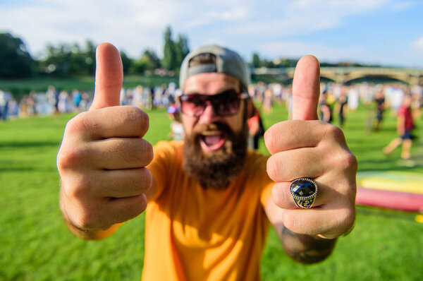 Мужчина бородатый на фоне толпы на берегу реки. Забронируйте билет сейчас на летний фестиваль. Очень рекомендуем мероприятия из топ-списка. Хипстерский пикник или фестиваль. Мужчина веселый лицо показывает большой палец вверх
