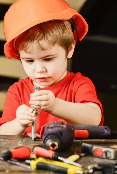 Kind mit Bollenhut, Helm, der mit Sechskantschrauben spielt, als Bauarbeiter oder Reparateur, Handarbeit. Kleinkind auf geschäftigem Gesicht spielt zu Hause in der Werkstatt mit Schrauben. Junge spielen als Handwerker. Bastelkonzept — Stockfoto