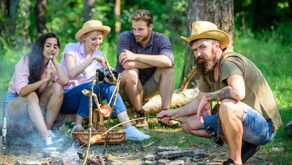 Amici gruppo turistico rilassante vicino al falò. Hipster arrostire salsiccia mentre gli amici si siedono sul log sharing impressione e guardare le foto sulla macchina fotografica. Turisti che fanno merenda con arrosto sul cibo fuoco — Foto Stock