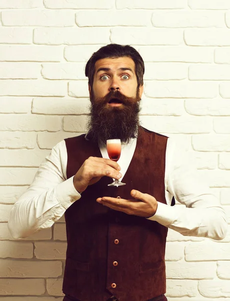handsome bearded man with long beard and mustache has stylish hair on surprised face holding glass of alcoholic shot in vintage suede leather waistcoat on white brick wall studio background.