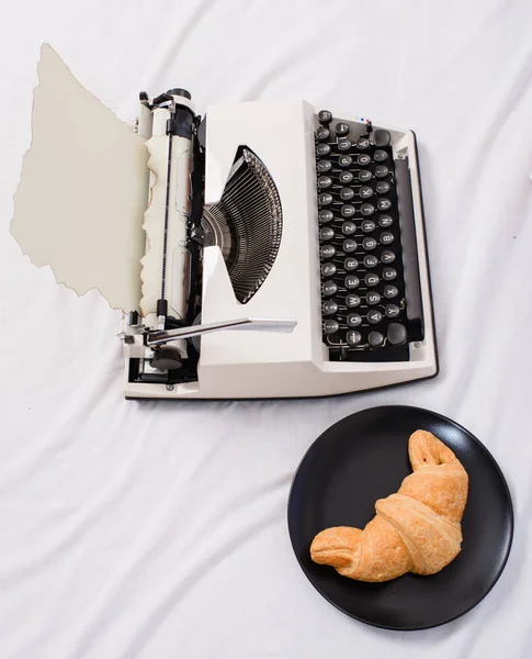 Máquina de escribir y delicioso plato de croissant ponen sábanas blancas. Beneficio ser escritor es cómodo lugar de trabajo inspirador. Crear atmósfera inspiradora antes de empezar a escribir una nueva página con la máquina de escribir — Foto de Stock