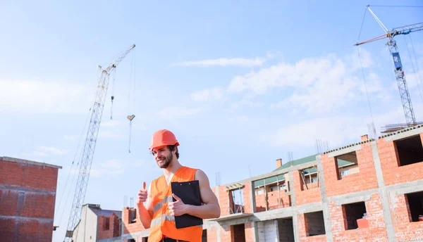 男子橙色背心和头盔工程施工现场 承包商按计划控制 控制施工过程 戴着防护头盔的家伙站在用红砖砌成的建筑物前 — 图库照片