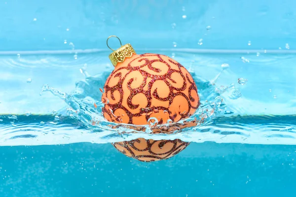 Holidays and vacation concept. Christmas decoration or toy for Christmas tree swim in pool. Festive decoration for Christmas tree, orange ball with glitter decor dropped into water, blue background