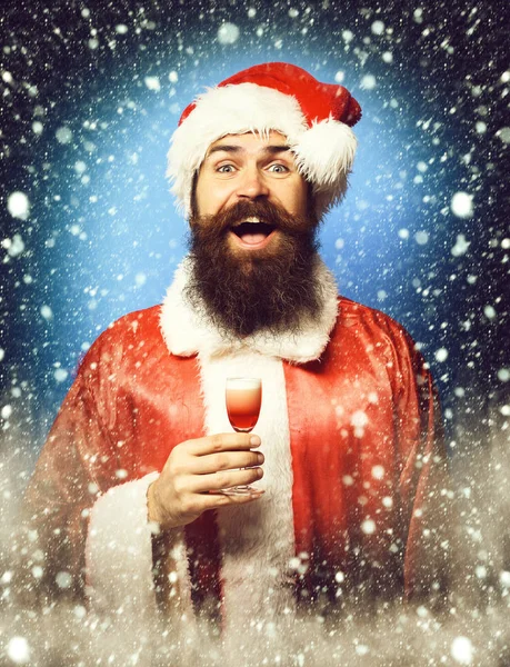 Guapo barbudo hombre de Santa Claus con barba larga en la cara sonriente sosteniendo vaso de trago alcohólico en navidad roja o suéter de Navidad y sombrero de año nuevo en fondo de estudio azul —  Fotos de Stock